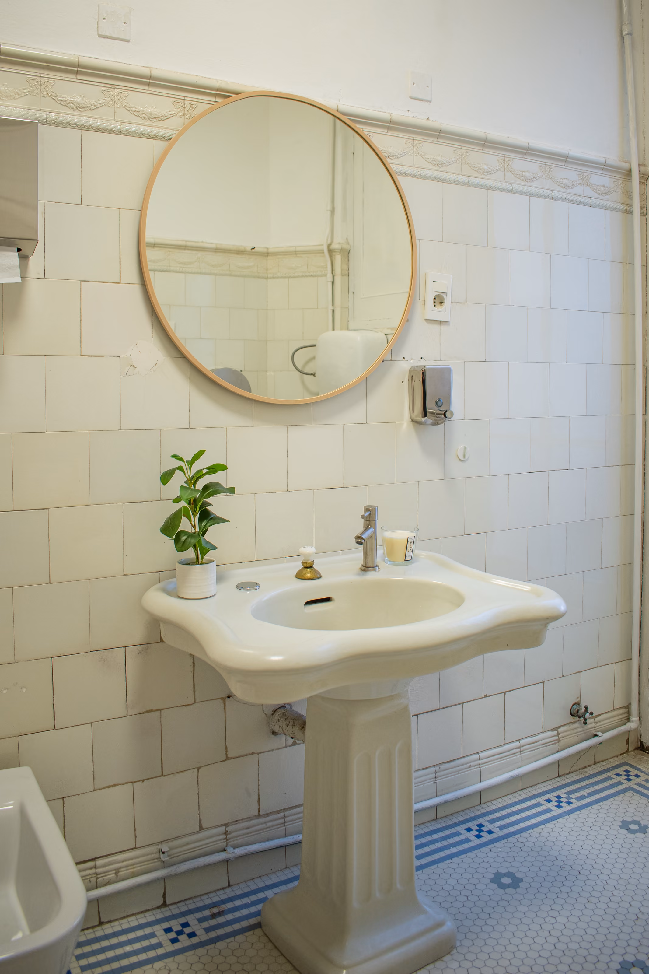Baño con lavatorio de pie, espejo redondo de marco claro, azulejos blancos y piso de mosaico con guarda azul.