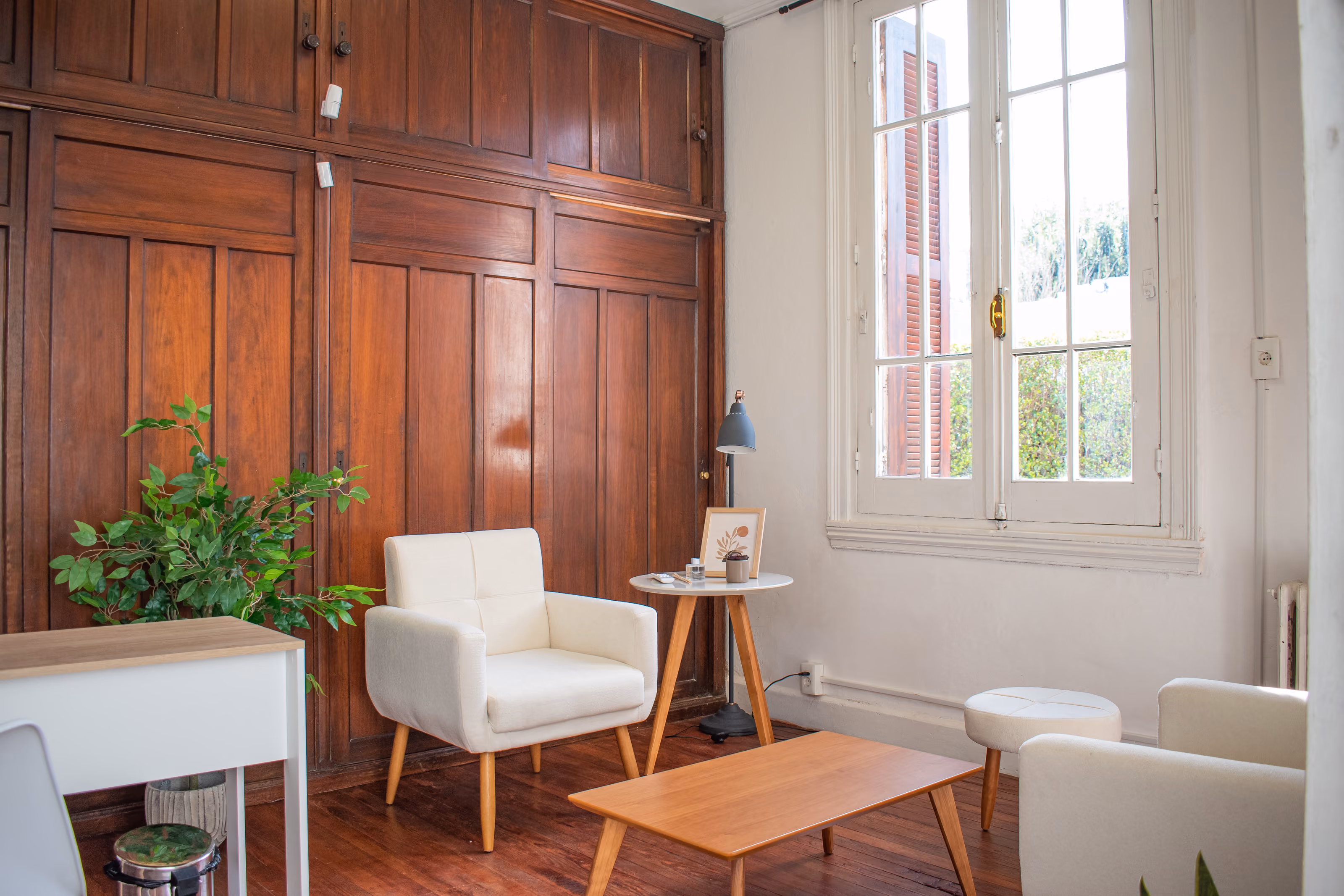 Consultorio 01: sillón blanco y mesa baja de madera contra una pared de madera original, junto a una ventana alta.