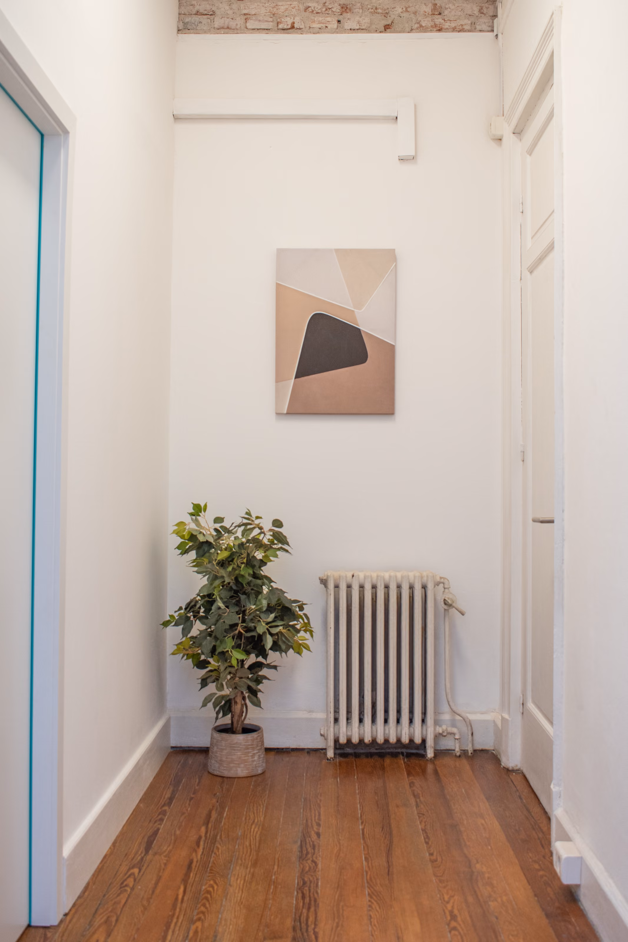Pasillo interior con paredes blancas, un cuadro de formas abstractas en tonos tierra, una planta y piso de madera original.
